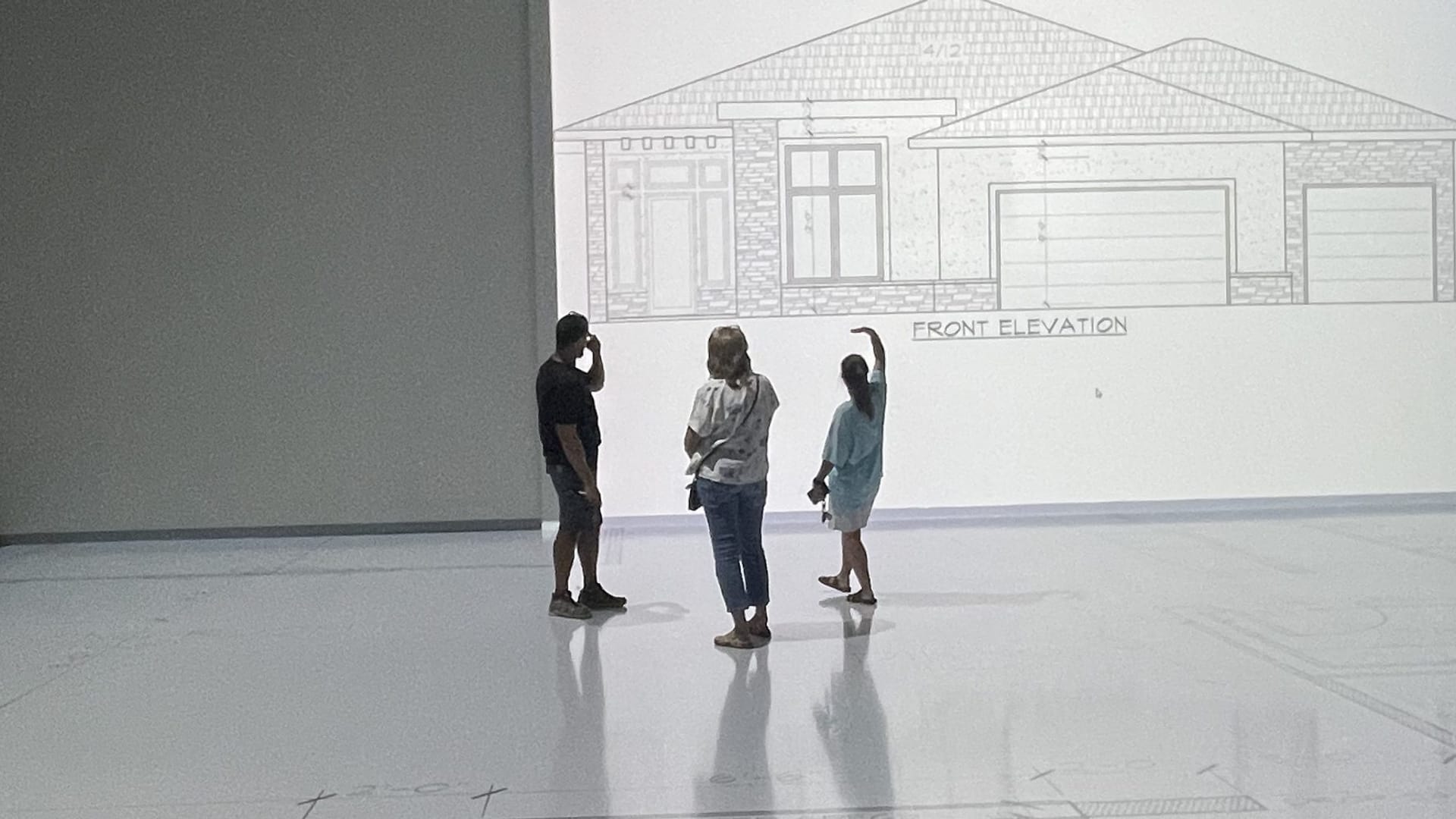 Clients walking a floor plan projected at full scale on the Walk Your Plans studio floor, with the home's front elevation projected on the wall